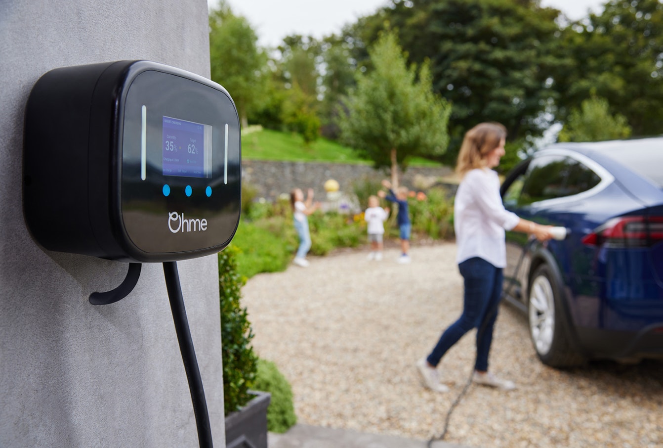 A close up of an Ohme home charging, with a mother plugging it into her car in the background and her 3 children throwing playing behind her