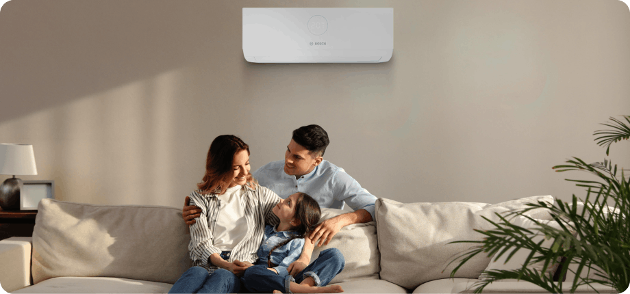 a smiling lady sat on a sofa on her phone, sun is shining in to the room through a window behind her 