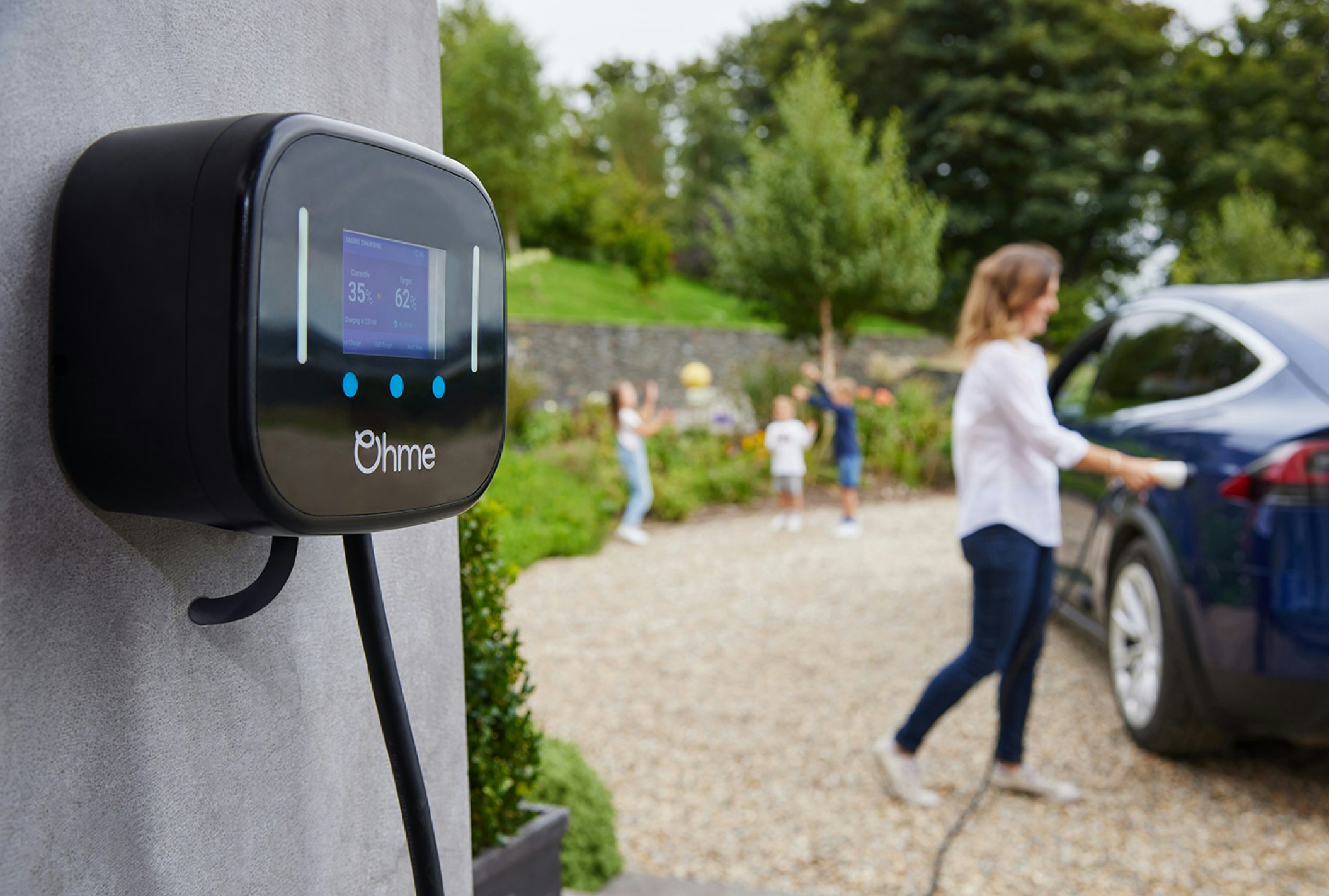 A close up of an Ohme home charging, with a mother plugging it into her car in the background and her 3 children throwing playing behind her