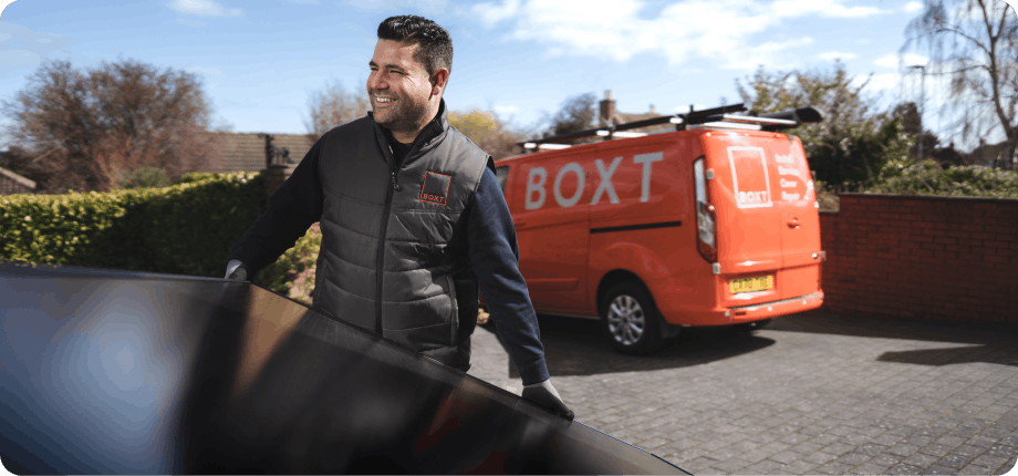 a smiling lady sat on a sofa on her phone, sun is shining in to the room through a window behind her 