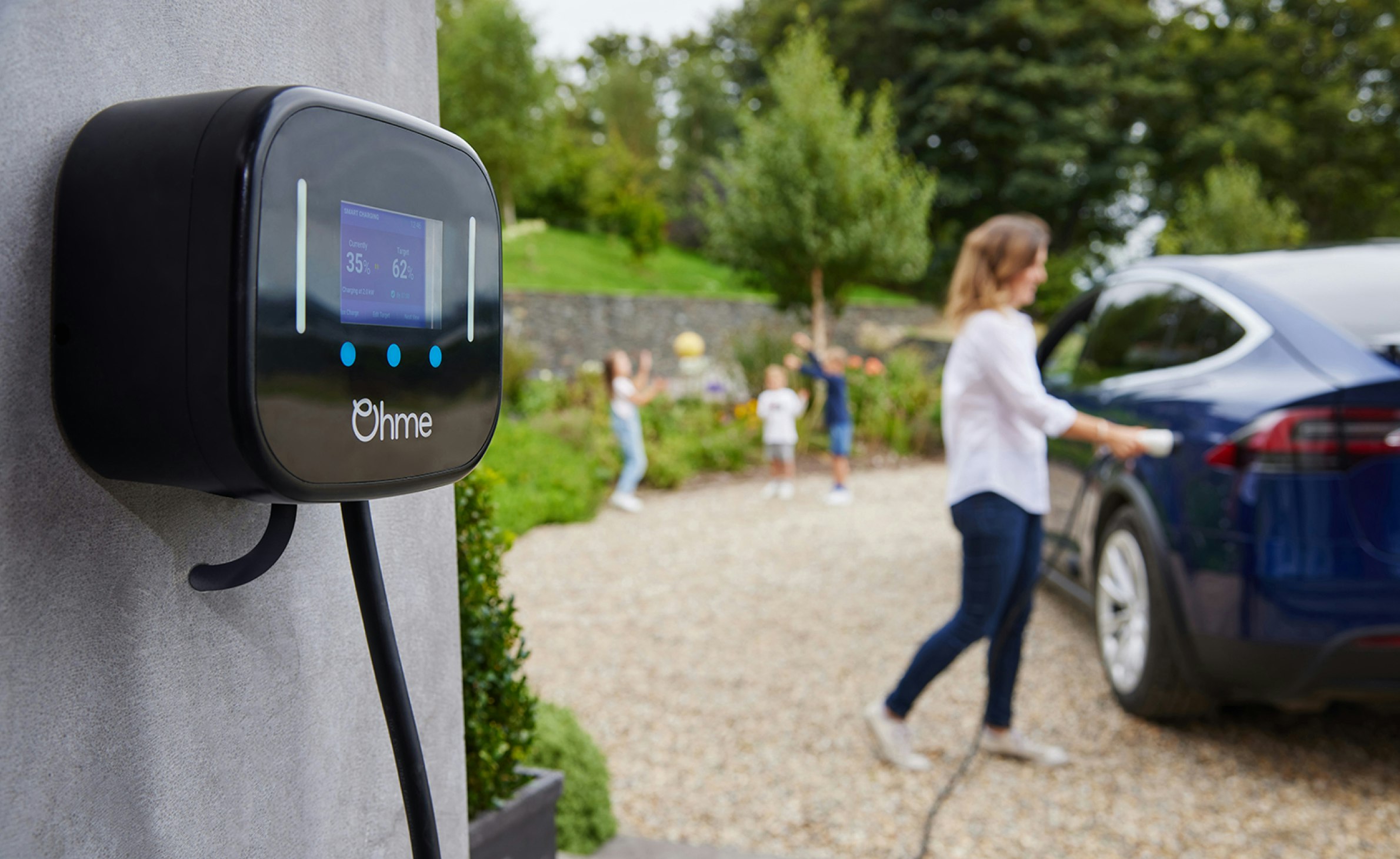 A close up of an Ohme home charging, with a mother plugging it into her car in the background and her 3 children throwing playing behind her